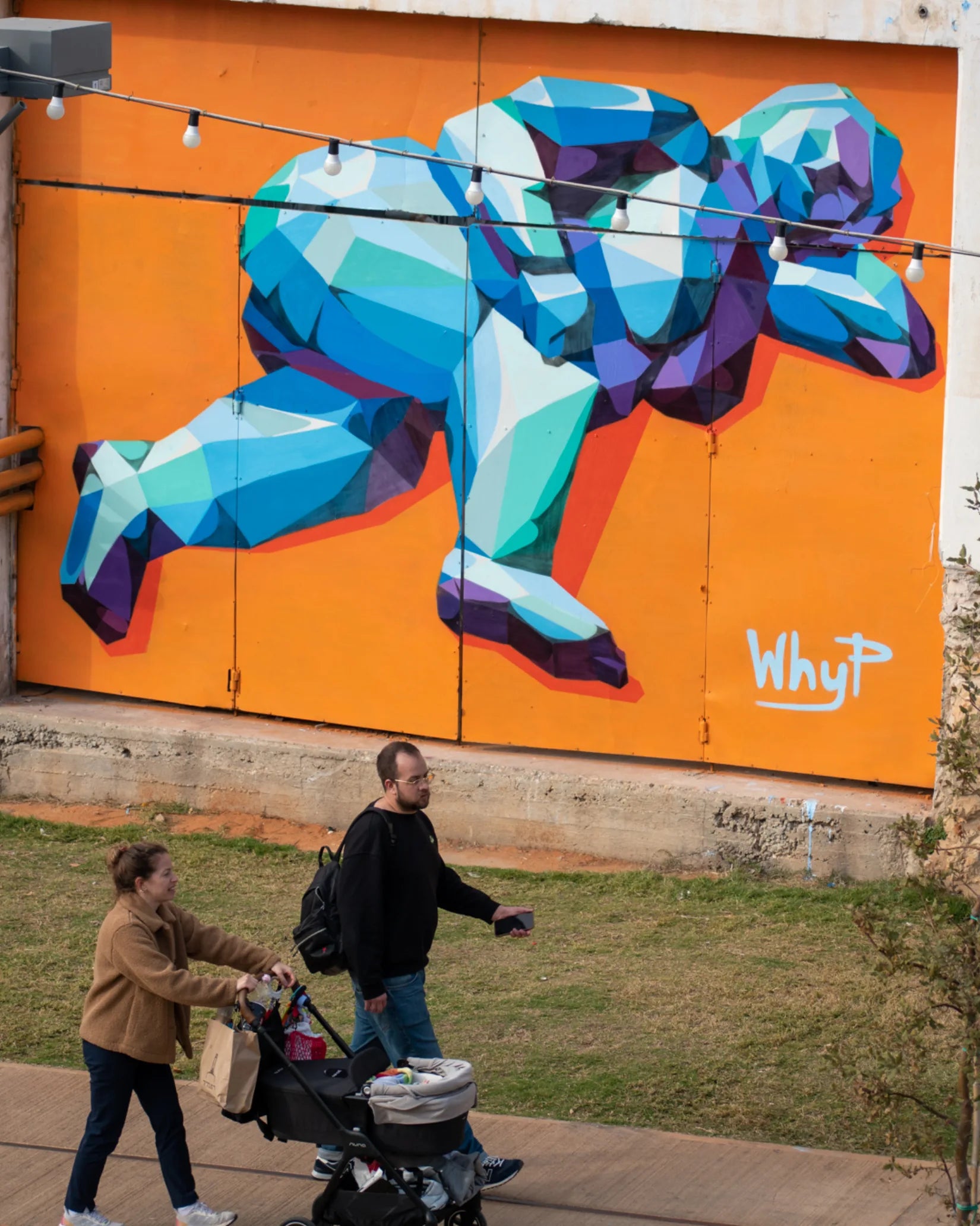 A blue and violet faceted figure painted by WHYP across an orange urban wall as people pass by